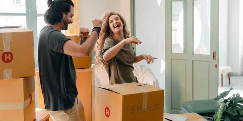 Cheerful laughing couple in casual clothes having fun and dancing together while unpacking carton boxes after moving into new contemporary apartment