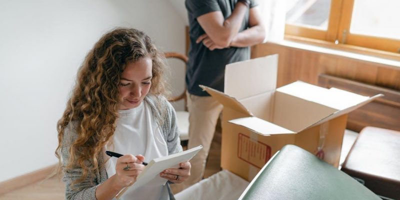 Pensive couple taking notes in bedroom near window while packing stuff in carton box to move into new apartment